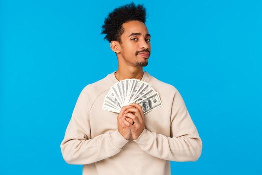 Suspicious and doubtful young pensive african-american rich guy holding big cash, lottery prize, have money and looking with disbelief or skeptical camera, unwilling share, standing blue background.