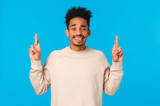 Nervous timid and cute african-american guy with afro haircut scared telling parents something huge, pointing fingers up, cringe and smiling with anxious expression, feeling alarmed, blue background.