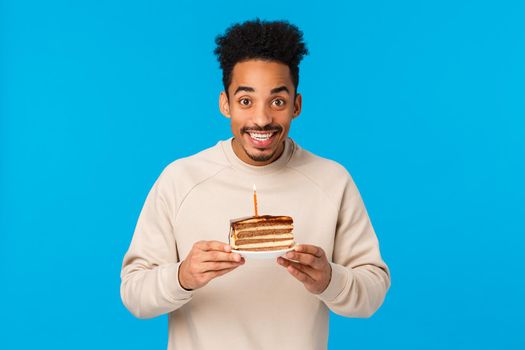 Cheerful and excited happy smiling african american b-day guy celebrating birthday, holding piece cake with candle, making wish ancitipating good year, standing blue background.