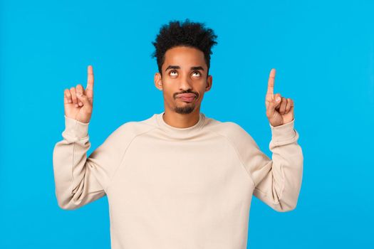 Guy got ugly sweater on christmas, feeling disappointed and upset. Handsome african american hipster guy with afro haircut, smirk skeptical and looking pointing up unsatisfied, blue background.