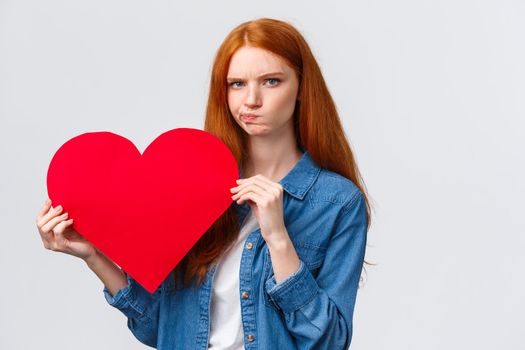 Serious-looking troubled, unsure cute redhead girl thinking what gift buy in addition to valentines day big red heart card, plan romantic date, pouting and frowning camera perplexed.