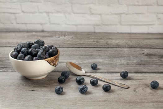 Small bowl full of blueberries, with more spilled next to spoon on gray wood desk.