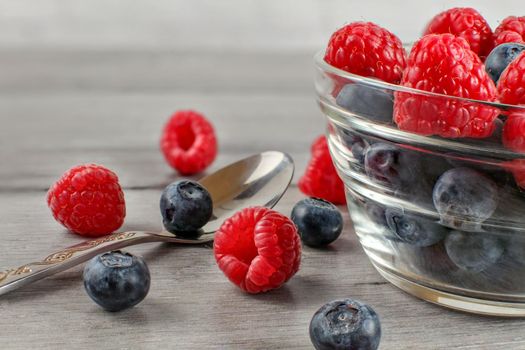 Closeup photo of bowl of blueberries and raspberries mix, some spilled on the gray wood desk and silver spoon next to it.