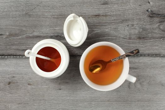 Tabletop view on white porcelain cup with hot tea and silver spoon, honey pot next to it, on gray wood desk.