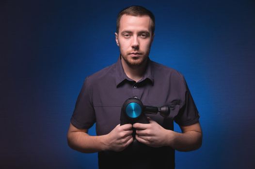 Portrait of a confident professional masseur with a percussion massager in his hands. Low key, blue background. Shock wave massage.