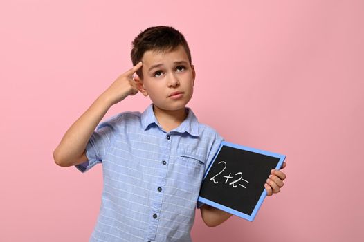 A schoolboy with a blackboard in his hand holds a finger to his temple and with a thoughtful look solves an arithmetic task, standing on a pink background with a place for text. Back to school. Concepts