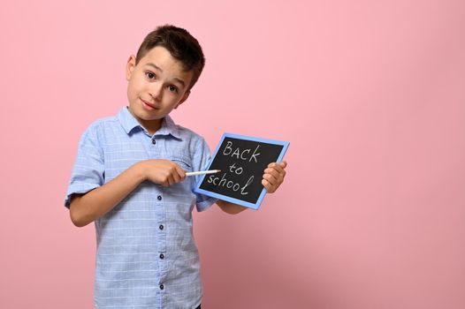 Smiling school boy holding a chalkboard with lettering Back To School and pointing with a pencil on it, posing over pink background with space for text. Concepts