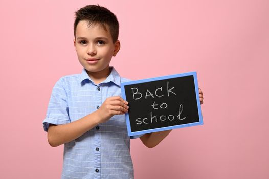 An adorable schoolboy holding a chalkboard with chalk lettering ,Back to school, isolated over pink background with space for text