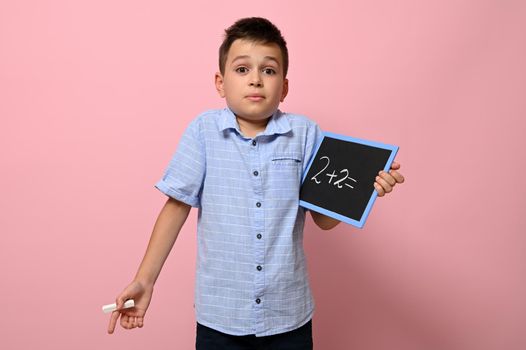 A cute schoolboy holds a blackboard in his hand with an arithmetic problem and with surprise and questions look at the camera while standing on a pink background with copy space
