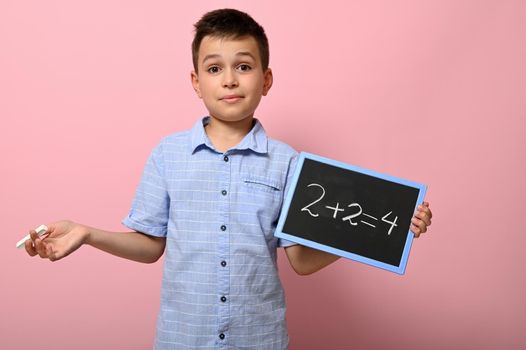 An adorable boy, elementary school student holds chalk and blackboard and solves math problem. Pink background space for text. Back to school. Concepts