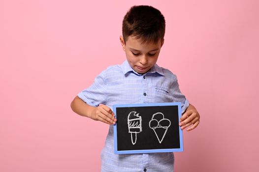 A school boy looking at a chalkboard in his hand with drawn ice cream against pink background with copy space. concepts