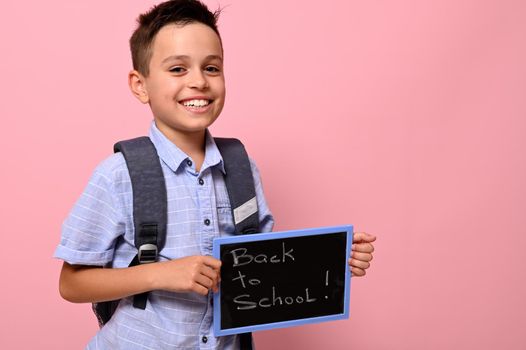 An adorable schoolboy with backpack holds chalkboard with chalk lettering ,Back to school, and cute smiles to camera, isolated over pink background with space for text