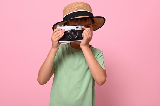 Cute schoolboy in green t-shirt and summer hat taking photo using an old vintage retro camera, isolated over pink background with copy space