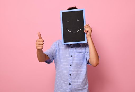 Unrecognizable school boy covers his face with a chalkboard with drawn smiling emoticons, expressing happiness and shows thumb up to the camera. Isolated over pink background with copy space