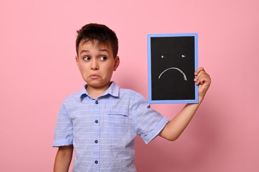 Isolated portrait on pink background of a sad schoolboy holding near his face a board with drawn sad emotion. Copy space.