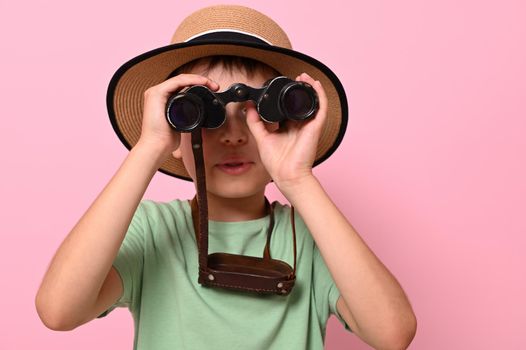 Close-up portrait of boy in summer straw hat looking through old vintage binoculars against pink background with copy space. Travel, tourism, summer, discovery and adventure concepts