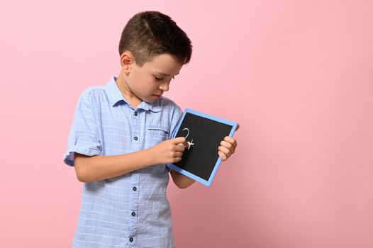 A school boy in blue shirt writes with chalk on a blackboard, isolated over pink background with copy space. Back to school. Concepts