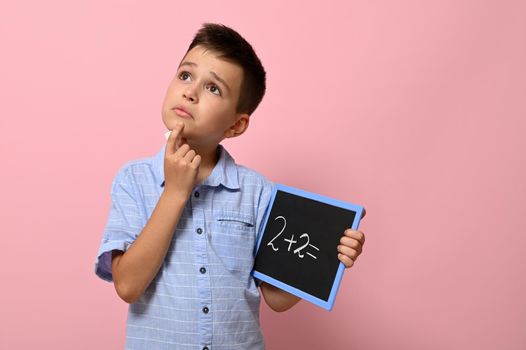 Cute schoolboy holding a blackboard with chalk writing on it, thoughtfully solves a math problem. Isolated on pink background with copy space. Back to school. Concepts