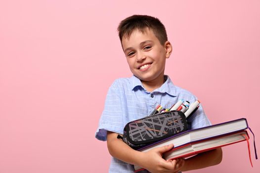Smiling school boy posing over pink background with books and pencil case full of stationery. Back to school. Concepts with facial emotions and space for text