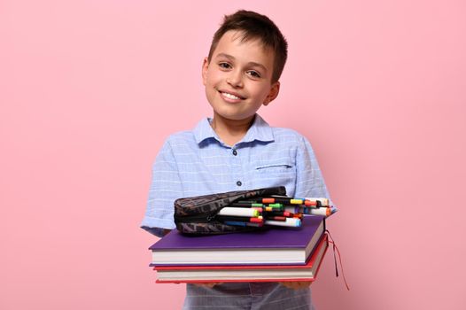 A smiling boy, student at school, holds books in front of him and pencil case with pens, felt-tip pens and markers falling out of him. Back to school concepts on pink background with copy space