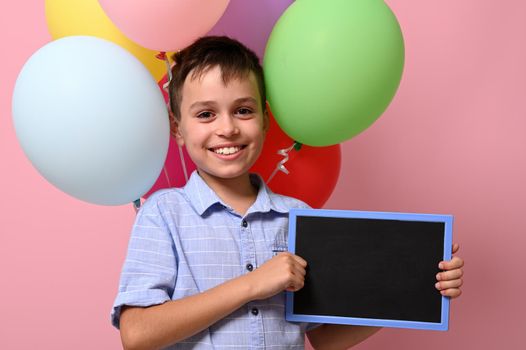 An adorable smiling schoolboy with empty blank chalkboard in his hand standing against multicolored balloons on pink background with copy space