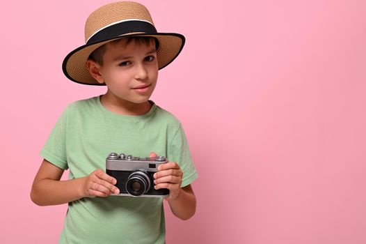 Handsome boy in summer hat holding a vintage retro camera, looks at the camera posing against pink background with space for text