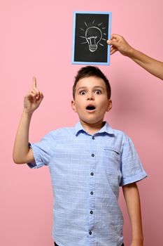 Concepts. Chalkboard with drawn lamp, idea symbol, and schoolboy looking pensively with forefinger pointing on a pink background with copy space. Emotions on backgrounds. Concepts.