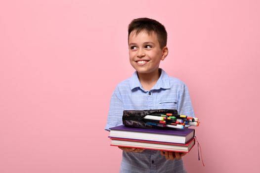 A charming boy, a student at school, holds books in front of him and pencil case with pens, felt-tip pens and markers falling out of him. Schoolboy cute smiles looking to the side on pink background