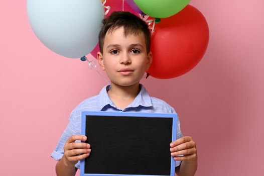 A handsome boy with blank chalkboard in his hand standing against multicolored baloons on pink background with copy space