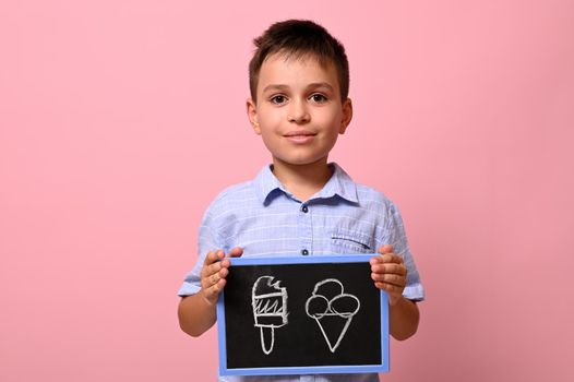 Isolated portrait on pink background of a handsome schoolboy holding a blackboard with drawn ice cream. Concepts.