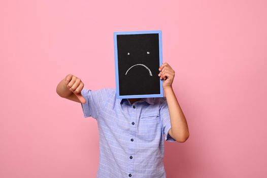 A school boy covers his face with a chalkboard with drawn smiling emoticons, expressing sadness and shows thumb down to the camera. Isolated over pink background with copy space