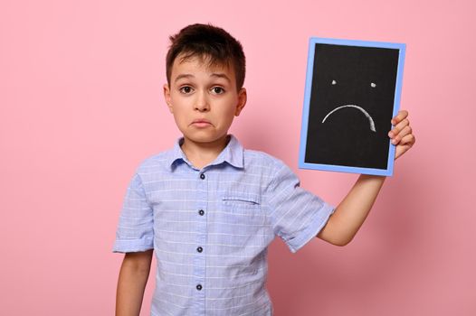 A frustrated boy in a blue shirt holds a blackboard near his face with a painted emoticon expressing frustration, sadness and disappointment