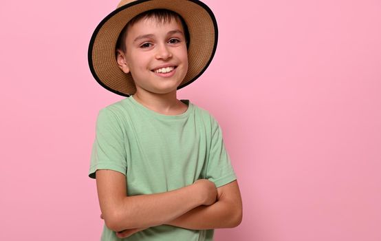 Happy and cheerful handsome boy in summer straw hat smiles with toothy smile ,standing with crossed arms on pink background while posing to camera. Kids emotions.