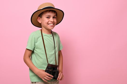 Handsome and cheerful schoolboy, tourist in summer outfit, with a retro vintage camera around the neck smiles looking at the camera while posing over pink background with copy space. Tourism concepts