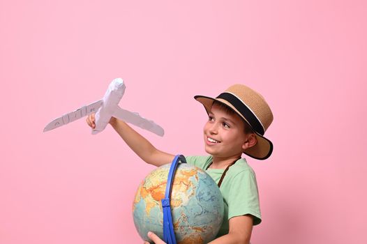 An adorable child boy plays with a paper airplane simulating a flight around the planet Earth. Concepts about travel, tourism, geography and aviation. Pink background with copy space