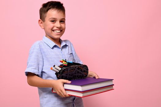 A smiling boy, student at school, holds books and pencil case in front of him. Back to school concepts on pink background with space for text