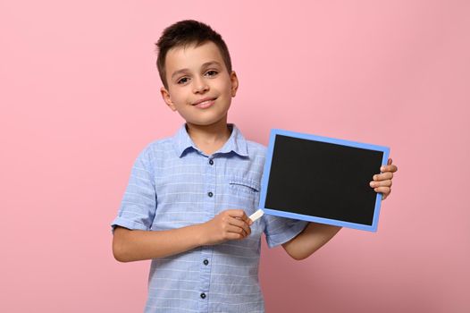 An adorable smiling boy pointing on an empty blank board with a chalk. Space for text on a chalkboard. Pink background with copy space. Concepts of back to school