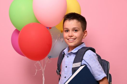 Concepts of happy back to school. Schoolboy with backpack holding book and multicolored balloons, cute smiling posing to camera over pink background with copy space