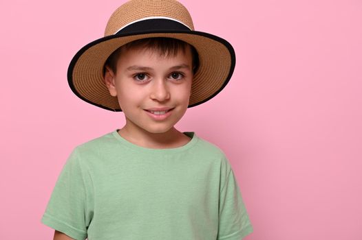 Close-up portrait of a handsome schoolboy in summer straw hat, looking at camera and standing isolated over pink background with space for text