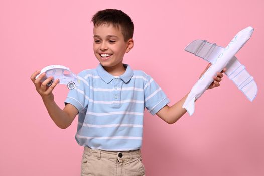 A handsome schoolboy holds a paper automobile and an airplane in his hands, choosing which transport is best for traveling. Benefits of land and air transport