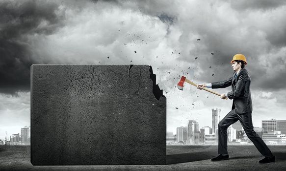 Young businessman in helmet breaking cement wall with axe