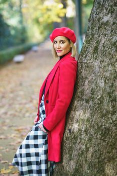 Beautiful middle-aged woman leaning on a tree trunk in a charming urban park. Female wearing coat, skirt and beret outdoors.