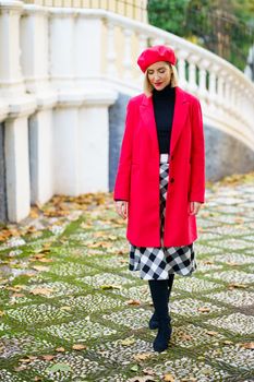 Beautiful middle-aged woman wearing red winter clothes walking through an urban park full of dry autumn leaves. Female wearing coat, skirt and beret outdoors.