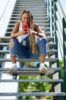Young black woman, with coloured braids, watching something very funny on her smartphone. Girl with a skateboard.