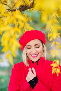 Happy middle-aged woman, smiling with her eyes closed among autumn leaves. Female wearing red coat, skirt and beret outdoors.