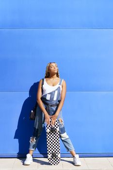 Black young girl dressed casual, wtih a skateboard on blue wall background.