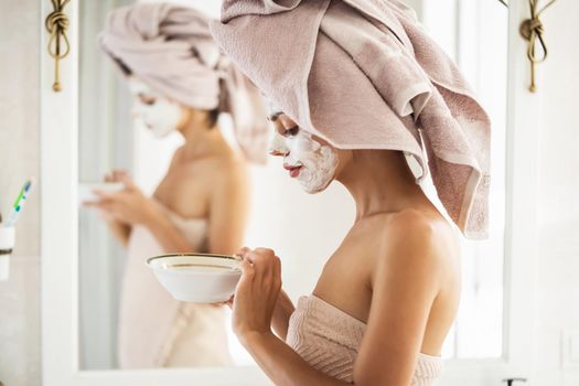 A young girl in a towel on her head and body stands near the mirror in the bathroom and applies a clay mask to her face, the woman takes care of her health and beauty.