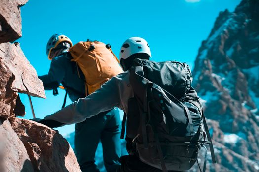 Two climbers hike to the top of the mountain with climbing, tourist equipment, men in helmets, with a rope and backpacks travel through the snow-capped mountains, rocky terrain.