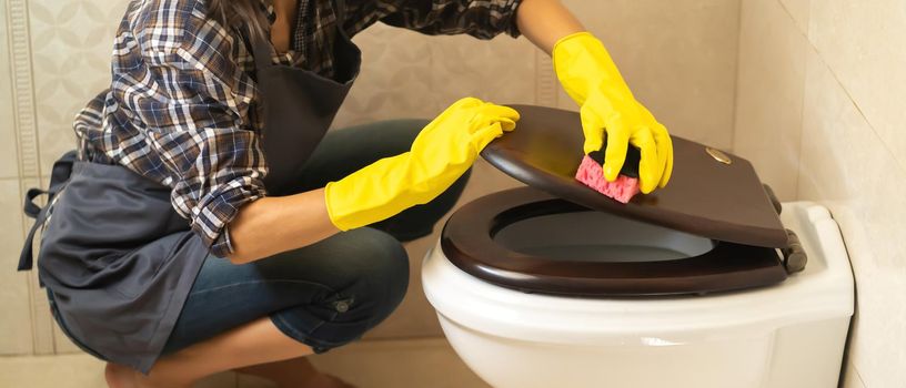 Female hands in yellow rubber gloves are holding a rag and wiping the toilet with detergent and disinfectant. The woman is cleaning the bathroom.