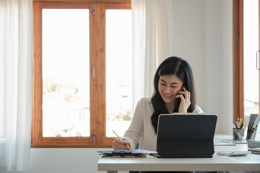 Happy business asian manageress working at her desk in the office taking a call on her mobile phone while writing notes on a notepad,